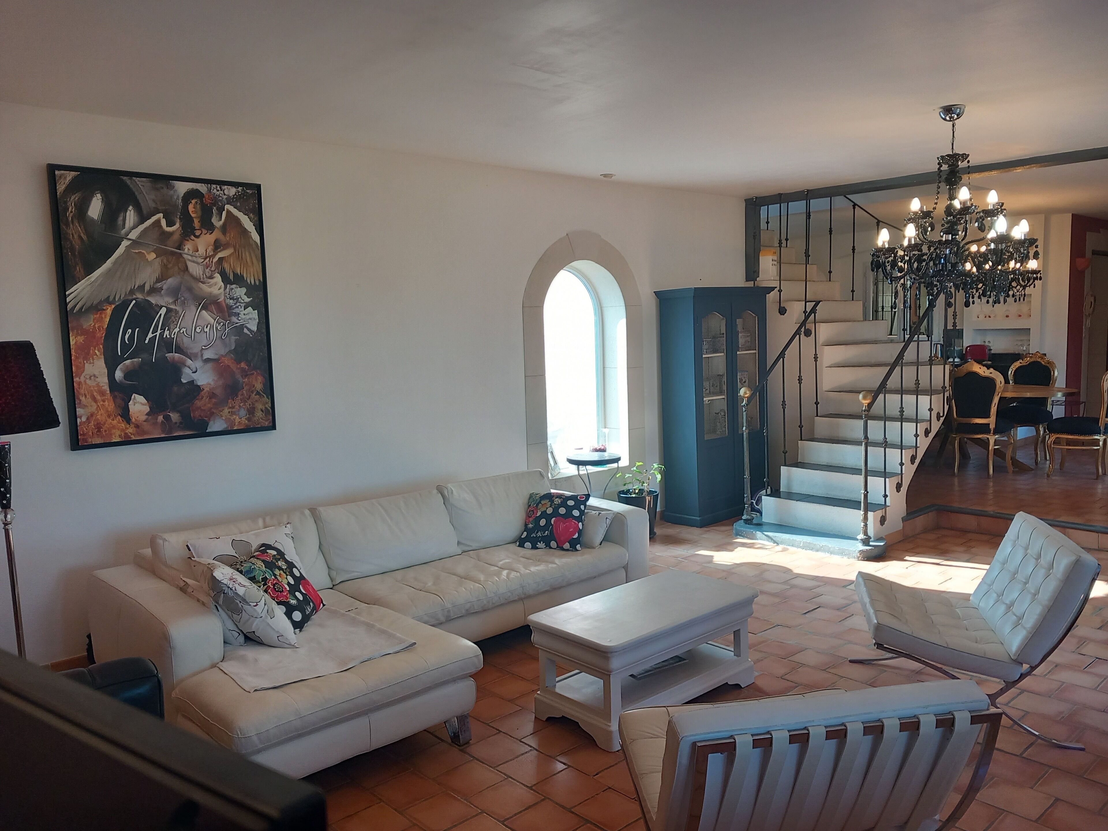 A spacious living room with a white sofa, a coffee table, and a blue armchair. A staircase leads to a dining area with a chandelier.