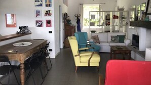Ein geräumiger Wohnbereich mit einem Esstisch, einem gelben Sessel, einem blauen Sessel und einem roten Sofa. Ein Bücherregal ist mit Büchern und Dekoration gefüllt.