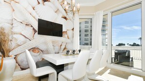 A modern dining area with a white table and chairs, a flat-screen TV mounted on a shell-patterned wall, and a large sliding glass door leading to a balcony with a view of buildings and trees. - An updated, modern star on the Gulf Coast shoreline! 🌟 (Destin)