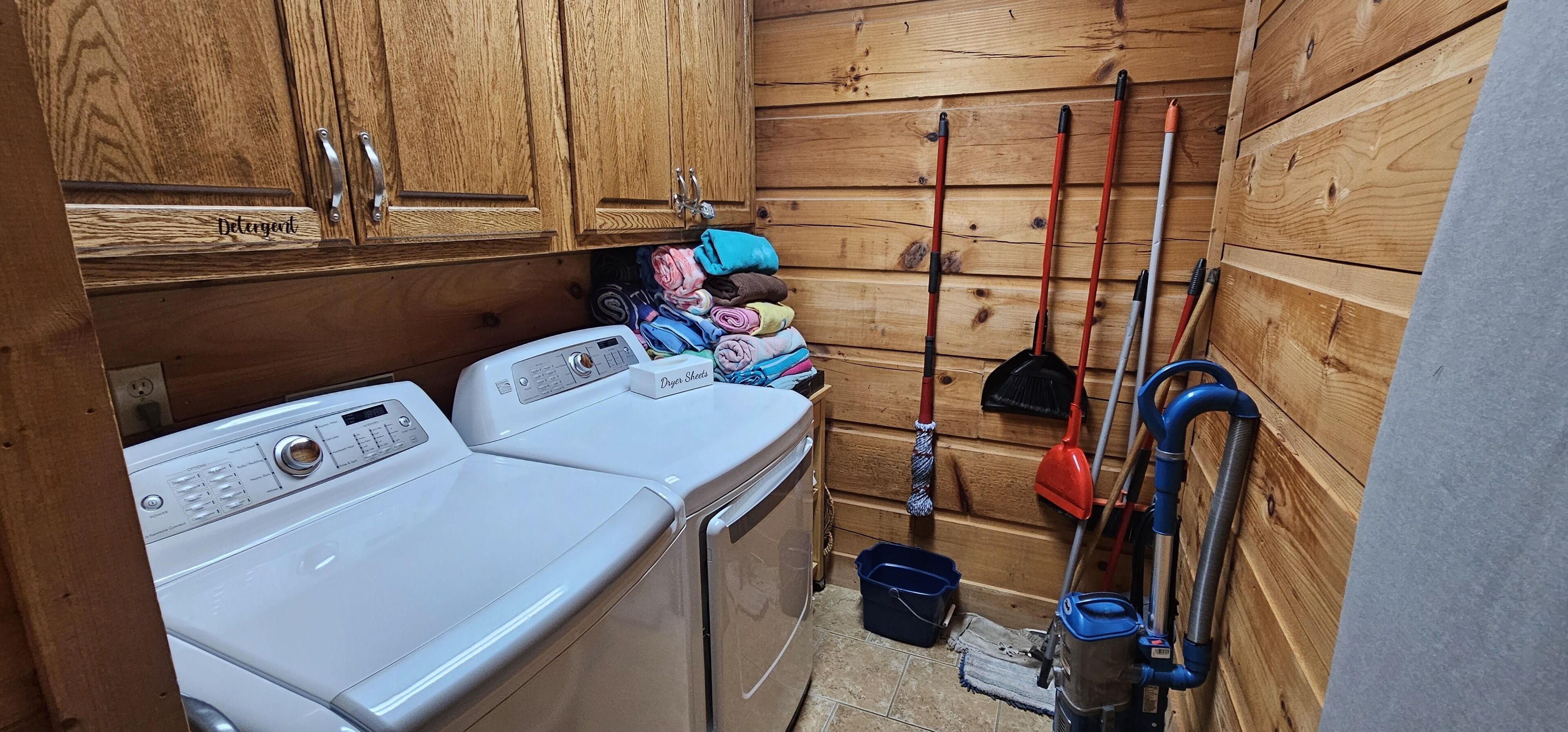 A laundry room with a washing machine, a dryer, a shelf with folded clothes, and cleaning tools.