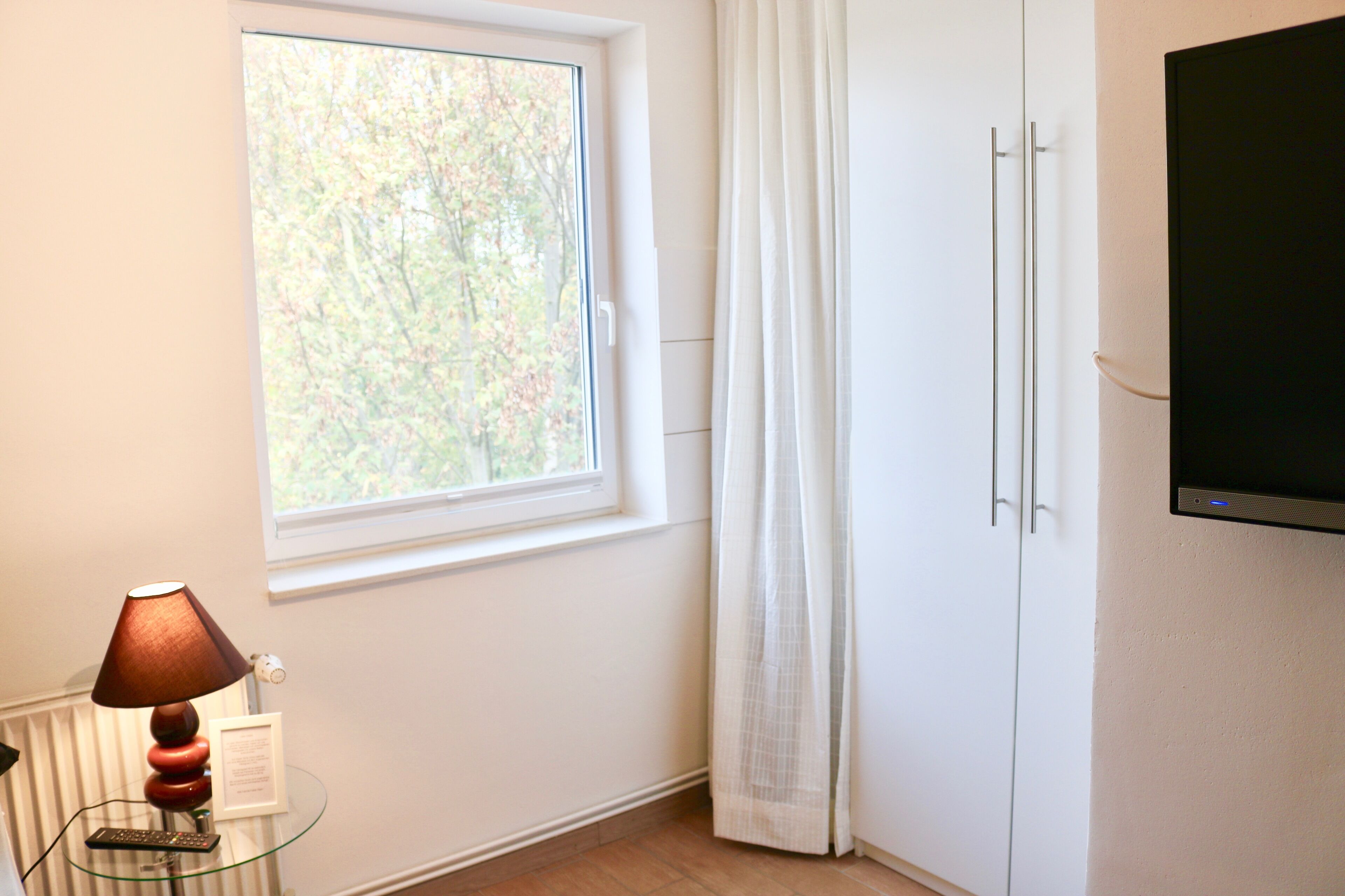 A room with a window, a lamp on a glass table, a white cabinet, and a flat-screen TV mounted on the wall.