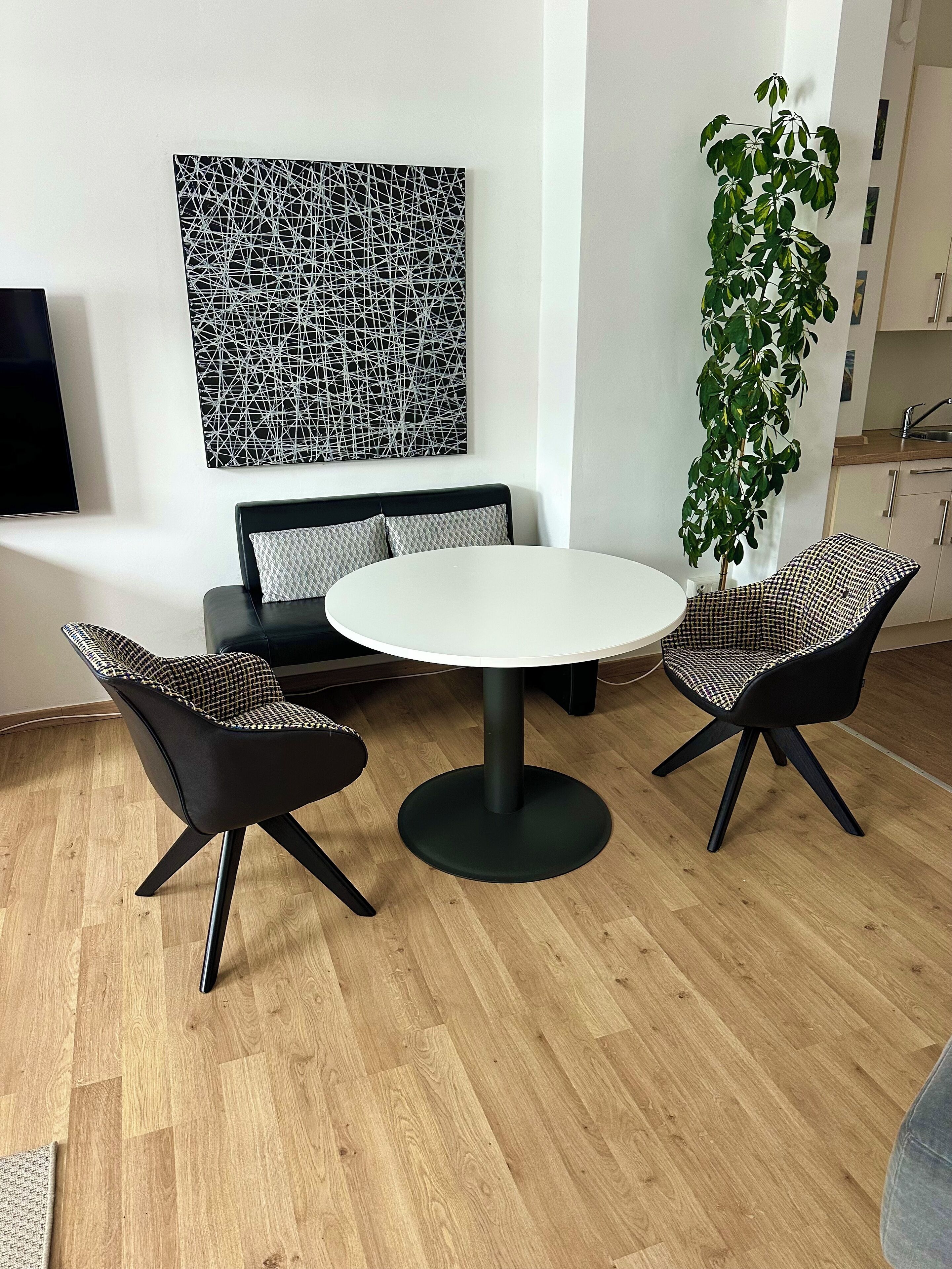 Ein modernes Wohnzimmer mit einem runden Tisch, zwei schwarz-weißen Schachbrettstühlen, einer Couch mit gemusterten Kissen, einem großen abstrakten Kunstwerk an der Wand und einer hohen Kühlpflanze.