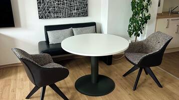 Ein modernes Wohnzimmer mit einem runden Tisch, zwei schwarz-weißen Schachbrettstühlen, einer Couch mit gemusterten Kissen, einem großen abstrakten Kunstwerk an der Wand und einer hohen Kühlpflanze.