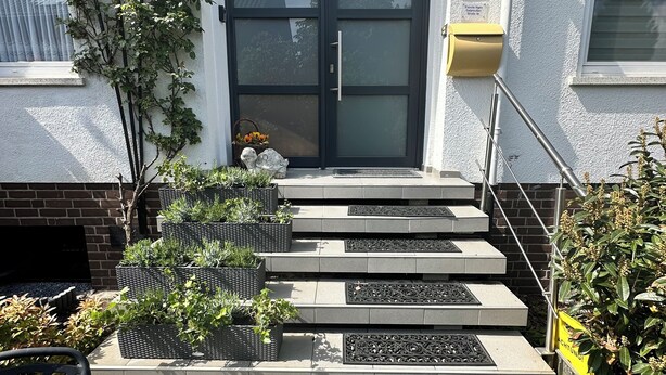 Une entrée moderne avec une porte noire, un petit espace jardin agrémenté de plantes et un banc en métal.