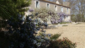 A rustic stone building with a tiled roof, surrounded by a gravel driveway and lush greenery including flowering shrubs and trees.