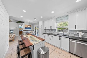 A modern kitchen with white cabinets, a central island with stools, a stainless steel sink, and a view of greenery through a window.