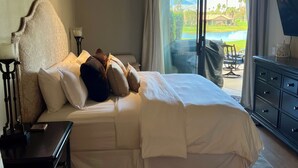 Habitación de hotel con una cama grande, una mesita de noche, un aparador y vistas a una piscina y follaje exterior.