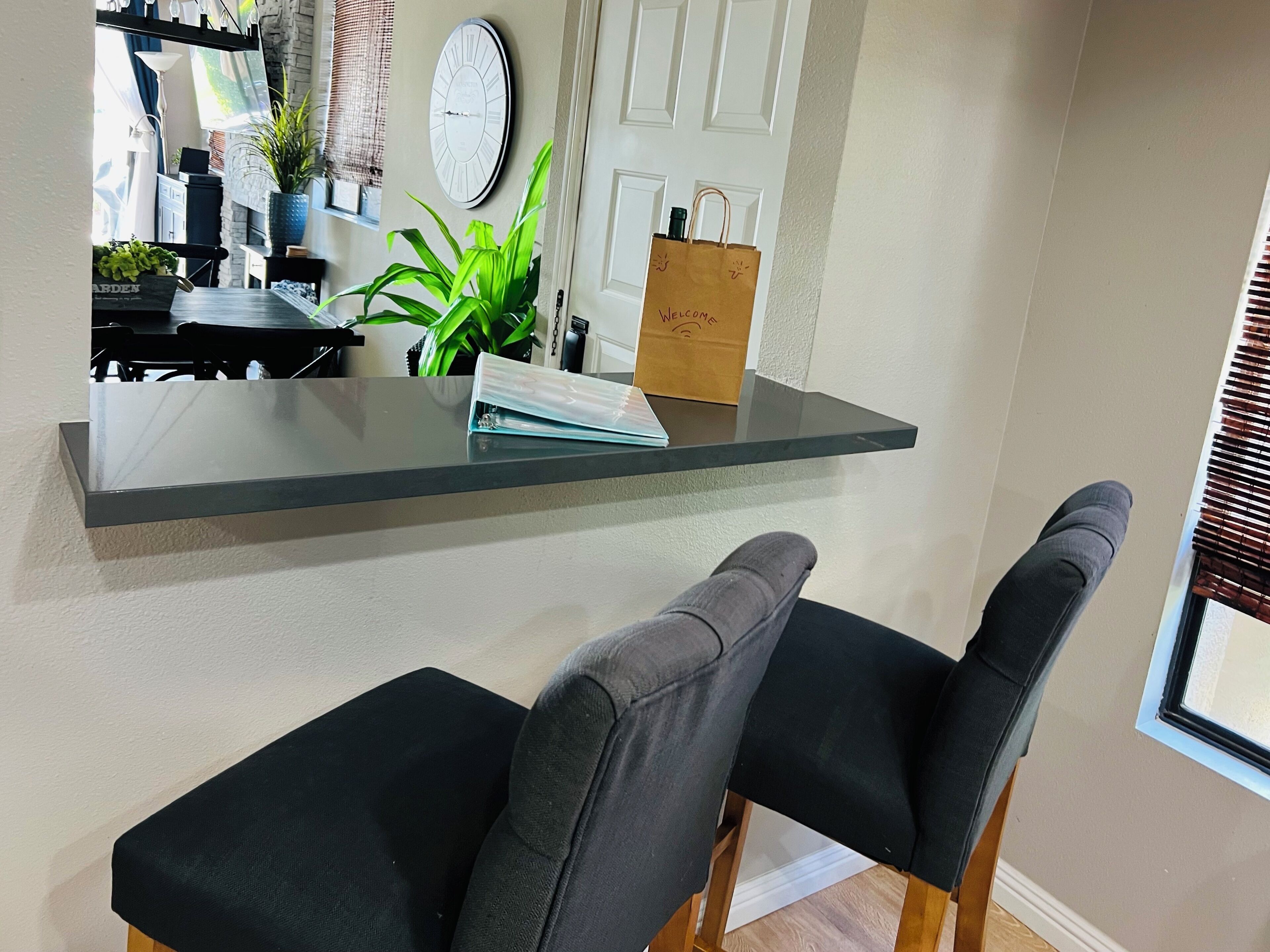A modern kitchen with a glass countertop, two black cushioned chairs, a potted plant, and a clock on the wall.