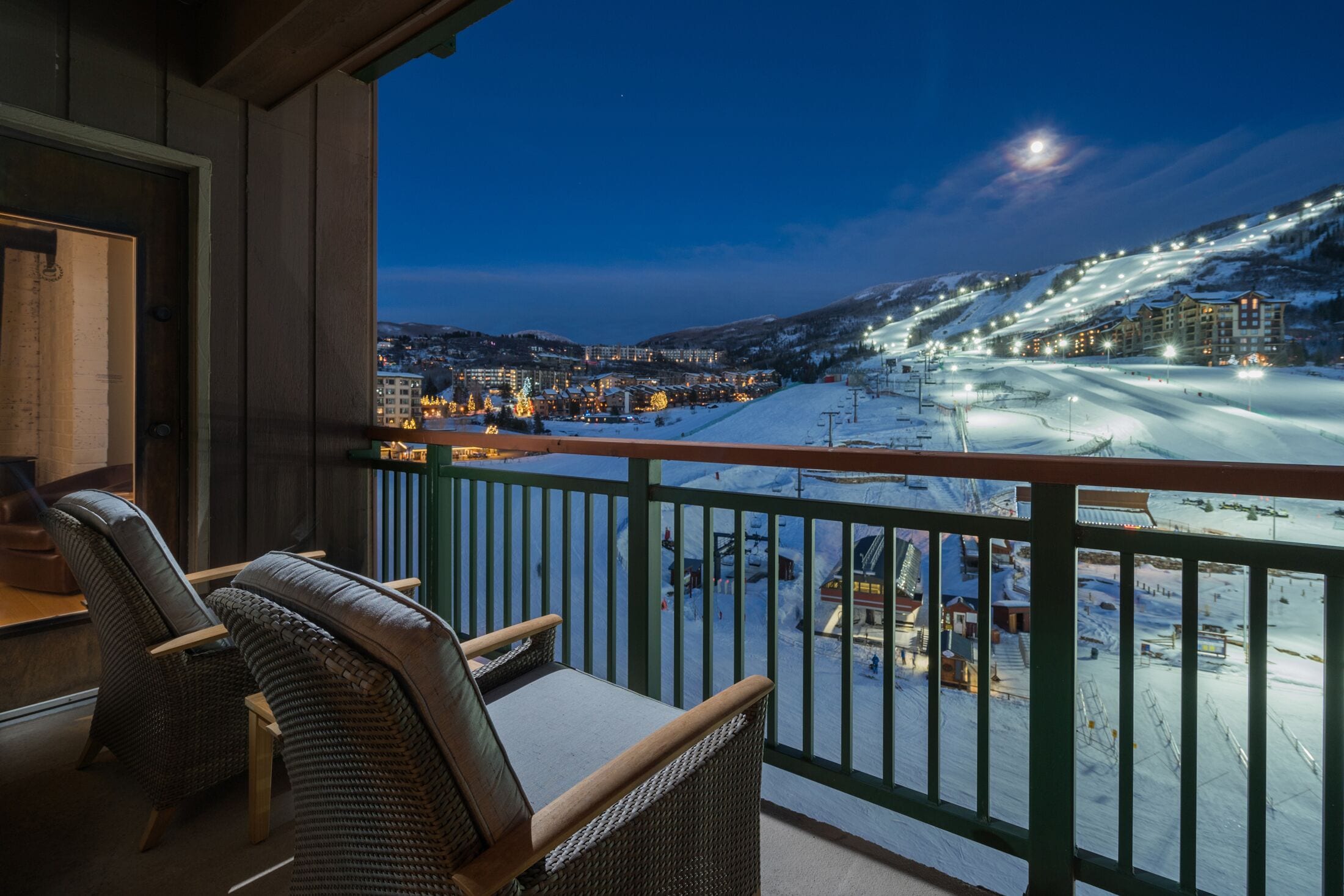A balcony with wicker chairs, overlooking a snow-covered ski slope and buildings at dusk.