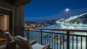 Un balcon avec des fauteuils en osier, donnant sur une piste de ski enneigée et sur des bâtiments à la tombée de la nuit.