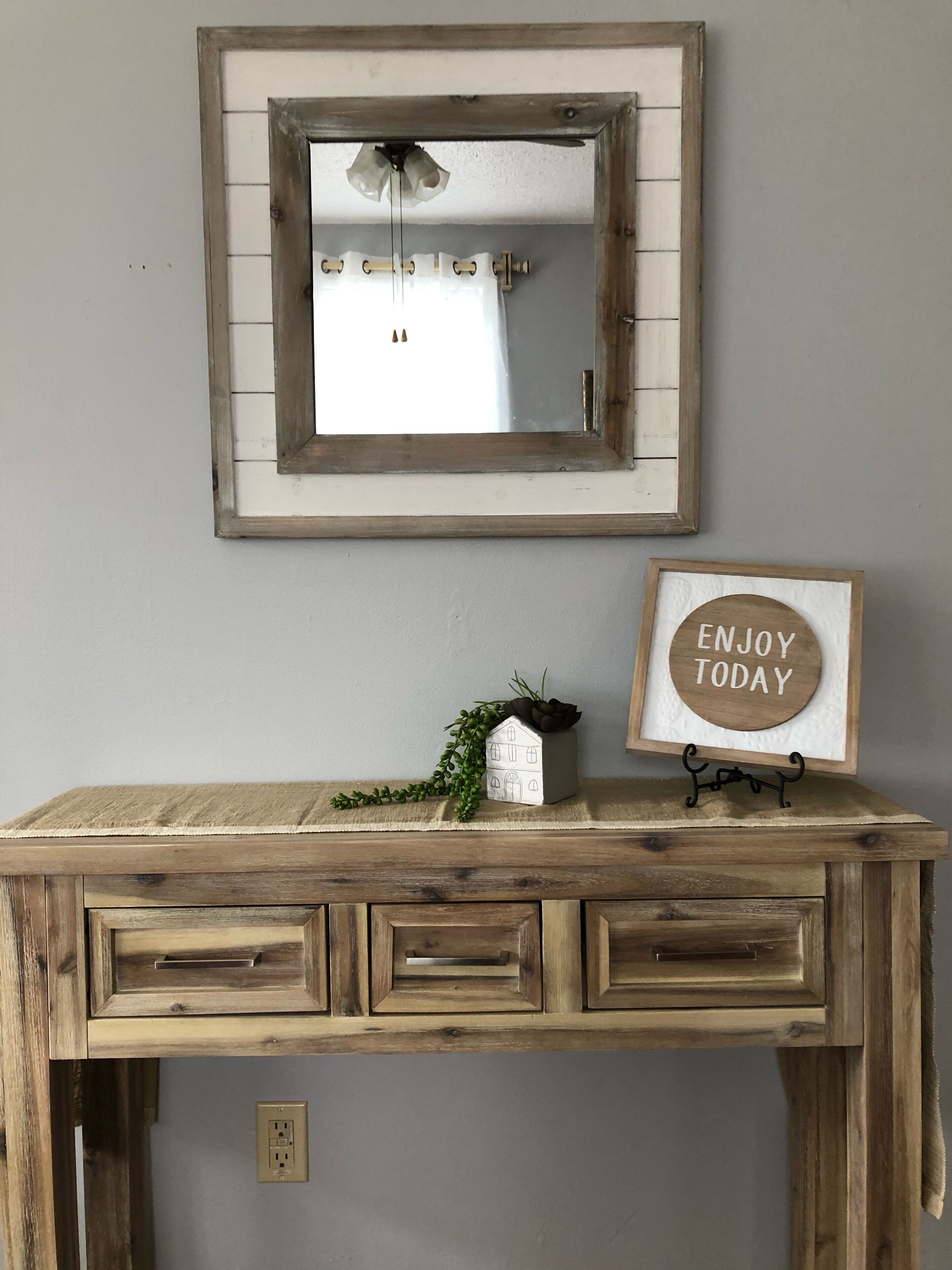 Un escritorio de madera rústico con tres cajones, una pequeña planta en maceta y un cartel enmarcado que dice 'ENJOY TODAY' montado en una pared sobre el escritorio.