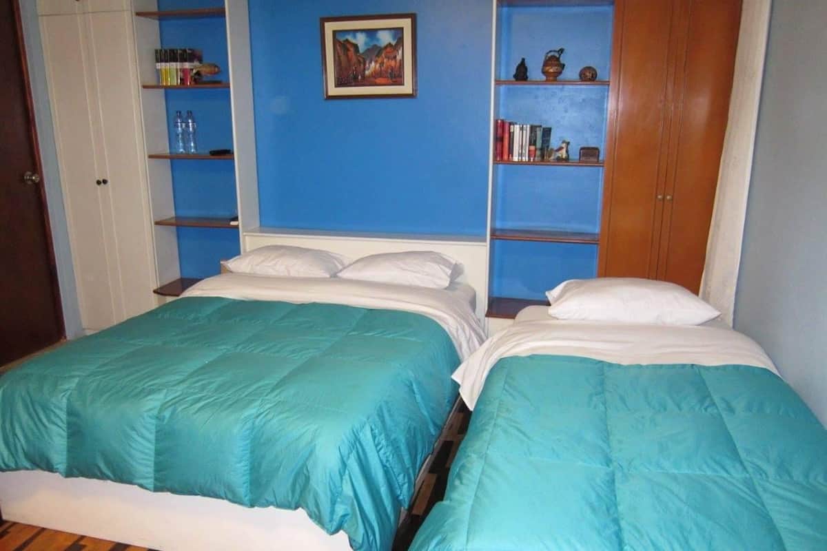 Une chambre avec deux lits, chacun agrémenté de draps de couleur turquoise, de têtes de lit en bois et d’oreillers blancs. On y trouve des étagères remplies de livres et d’objets décoratifs, ainsi qu’un tableau accroché au mur.
