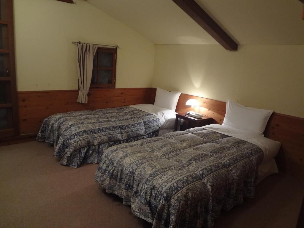 A room with two beds, a window with curtains, a nightstand with a lamp, and wooden beams on the ceiling.