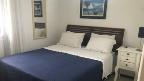A neatly made bed with a blue bedspread, a white pillow, and a wooden headboard. There is a small bedside table with a lamp and a framed picture on the wall above the bed. - Casa a 50 metros da praia da Armação (Florianópolis)