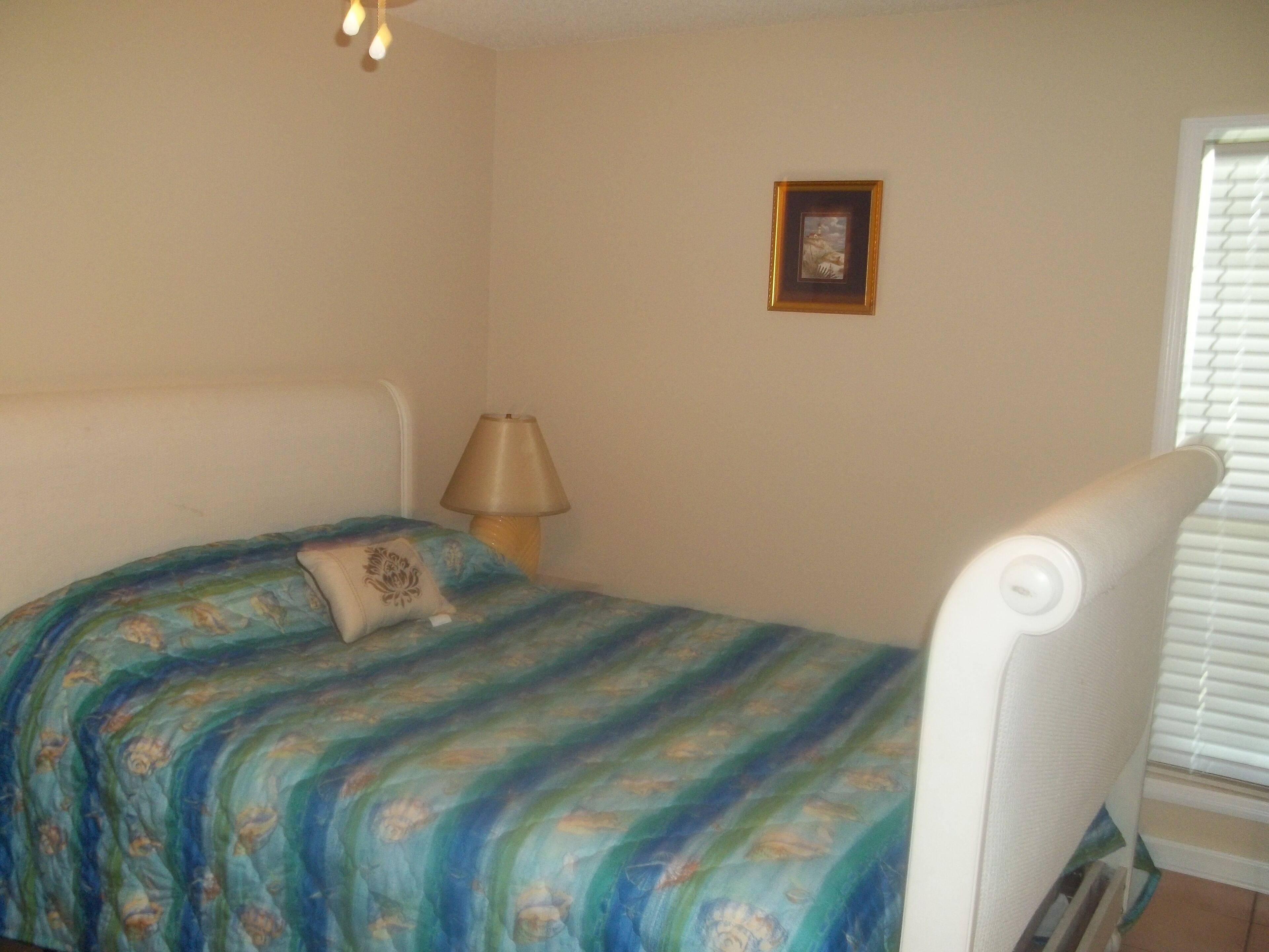 A bedroom with a bed, a bedside lamp, a picture on the wall, and a window with blinds.