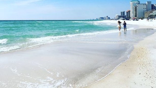 Ein Strand mit blauem Himmel, ruhigem Meer und einer Stadtkulisse im Hintergrund.