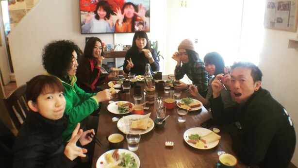 A group of people seated around a wooden dining table with various dishes, glasses, and a television mounted on the wall. - niseko KASHINOYA - Hostel (Kutchan)