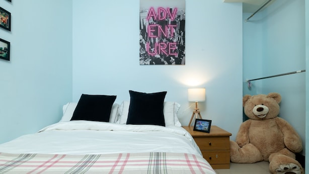 Ein Schlafzimmer mit einem Bett, Kissen, einem Nachttisch mit Lampe, einem Teddybären und einem Wandplakat.