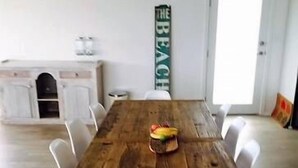 Une table à manger en bois, des chaises blanches, un panier de fruits et un panneau accroché au mur.