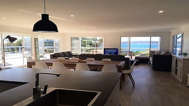 Une cuisine moderne dotée d’un îlot central, d’un coin repas avec une grande table et des chaises, et offrant une vue sur la mer grâce à de grandes fenêtres.