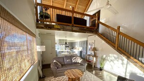 Un vaste espace de vie doté d’un haut plafond, d’un escalier en bois, d’une cuisine à l’arrière-plan et d’une grande fenêtre avec des stores.