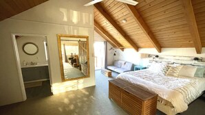 Une chambre avec un plafond en bois, un grand lit, un banc, un miroir et une salle de bain équipée d’un lavabo et d’un miroir.
