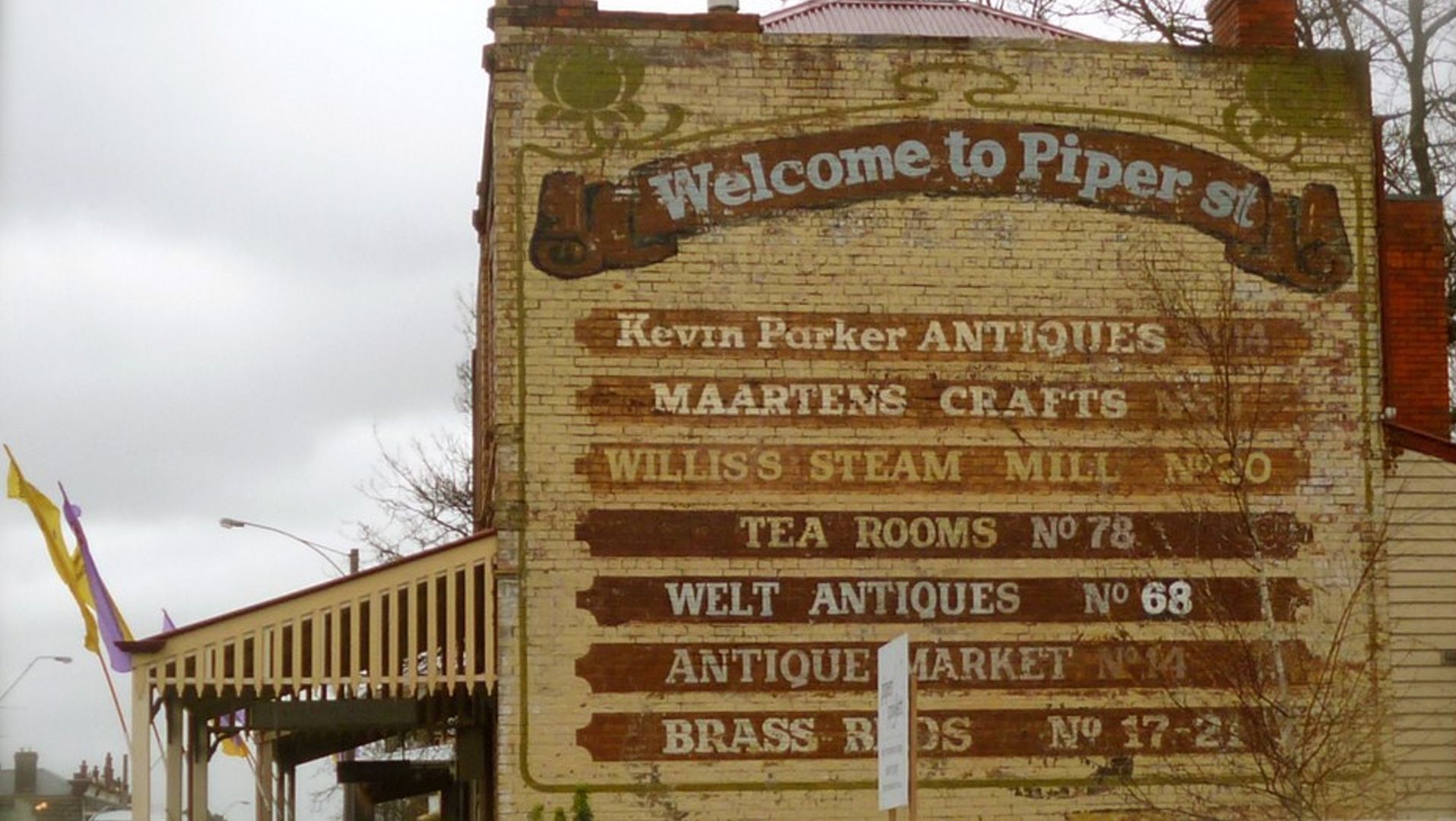Un mur de briques sur lequel est apposée une enseigne indiquant différents commerces, notamment Kevin Parker Antiques, Maartens Crafts, Williss Steam Mill, Tea Rooms No 78, Welt Antiques No 68, Antique Market No 14 et Brass Ribs No 17-2.