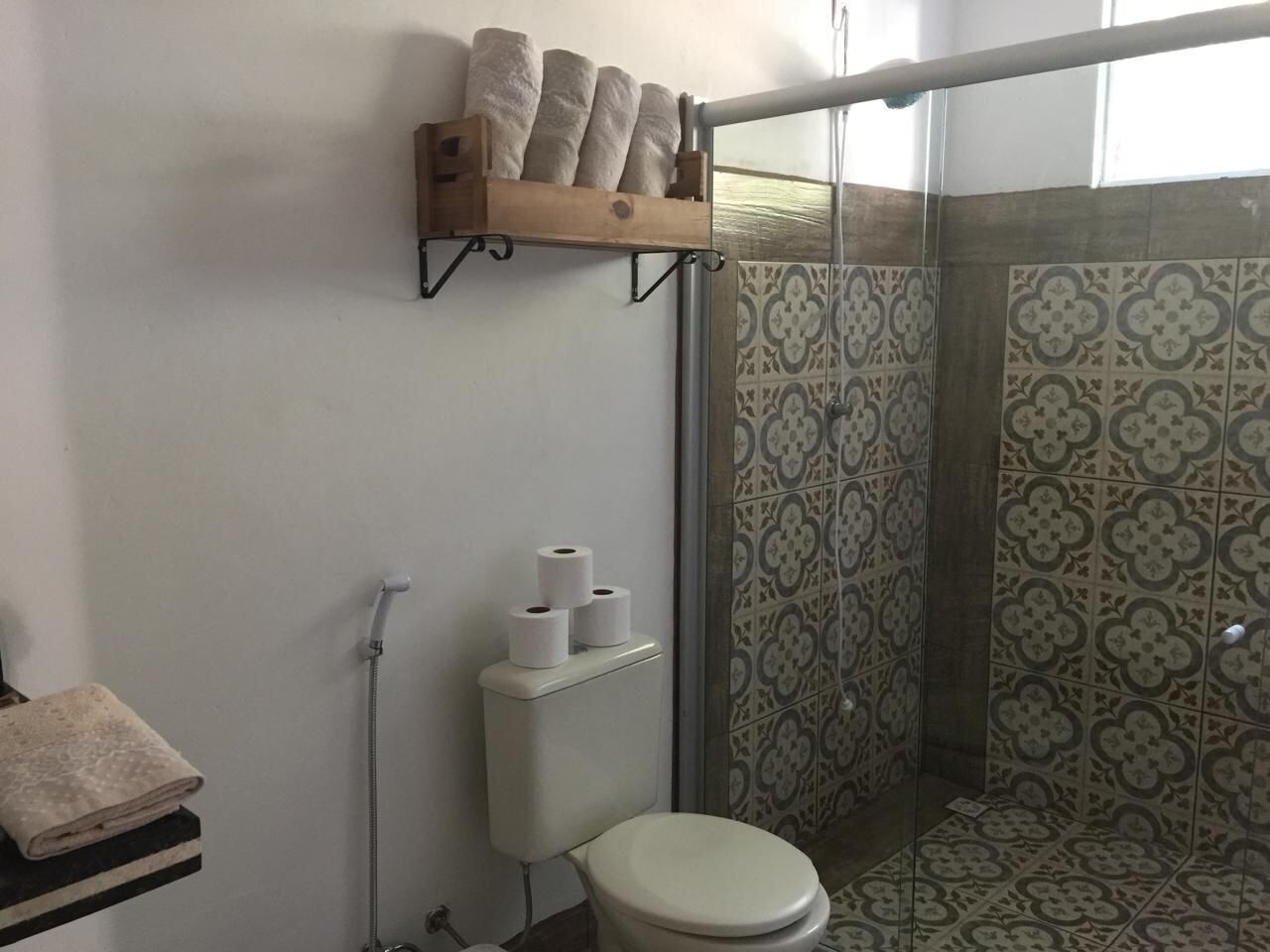 Casa de banho com sanita, chuveiro e azulejos estampados. Toalhas numa prateleira e papel higiénico na parede.