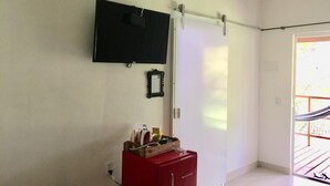 Una habitación con una televisión de pantalla plana montada en la pared, un pequeño mueble de madera con varios objetos, y una puerta que da a un espacio al aire libre.