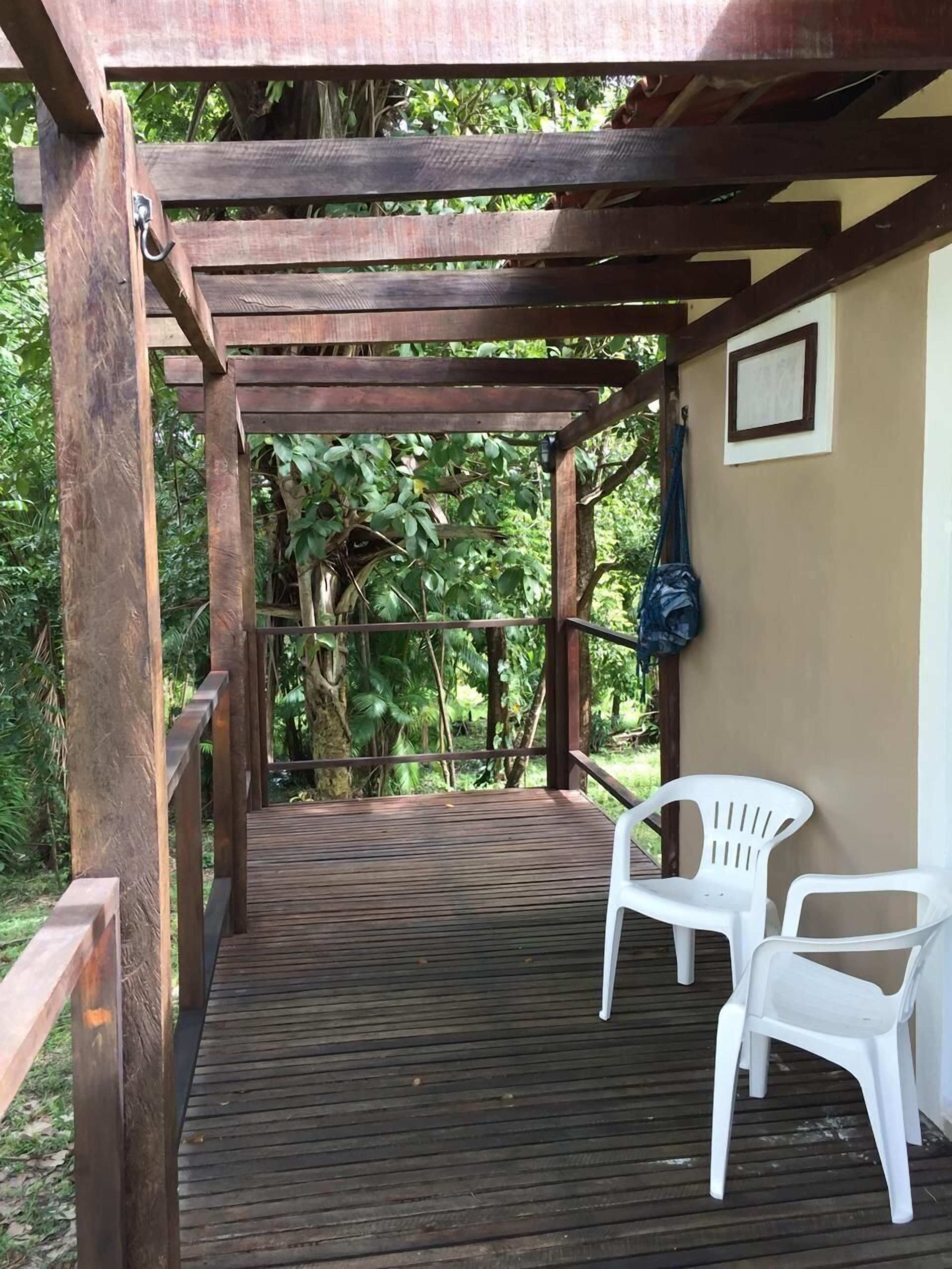 Una terraza de madera con dos sillas blancas de plástico, rodeada de follaje y una estructura de pérgola de madera.