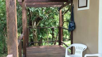 A wooden deck with two white plastic chairs, surrounded by greenery and a wooden pergola structure.