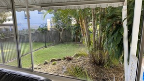 A view from a window of a room with a view of a backyard. The backyard has a grassy area, a black metal fence, and various plants including palm trees and a large bush. - ***Coastal Luxury*** Winter special 10% discount*** (Iluka)