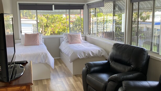 A room with two beds, a black leather sofa, a TV on a wooden stand, and large windows with a view of greenery outside. - ***Coastal Luxury*** Winter special 10% discount*** (Iluka)