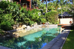 A clear pool surrounded by lush greenery and a building with a red roof. - Beautiful Ocean Views in the Heart of Port Douglas | 2 Bedroom Luxury Villa (Port Douglas)