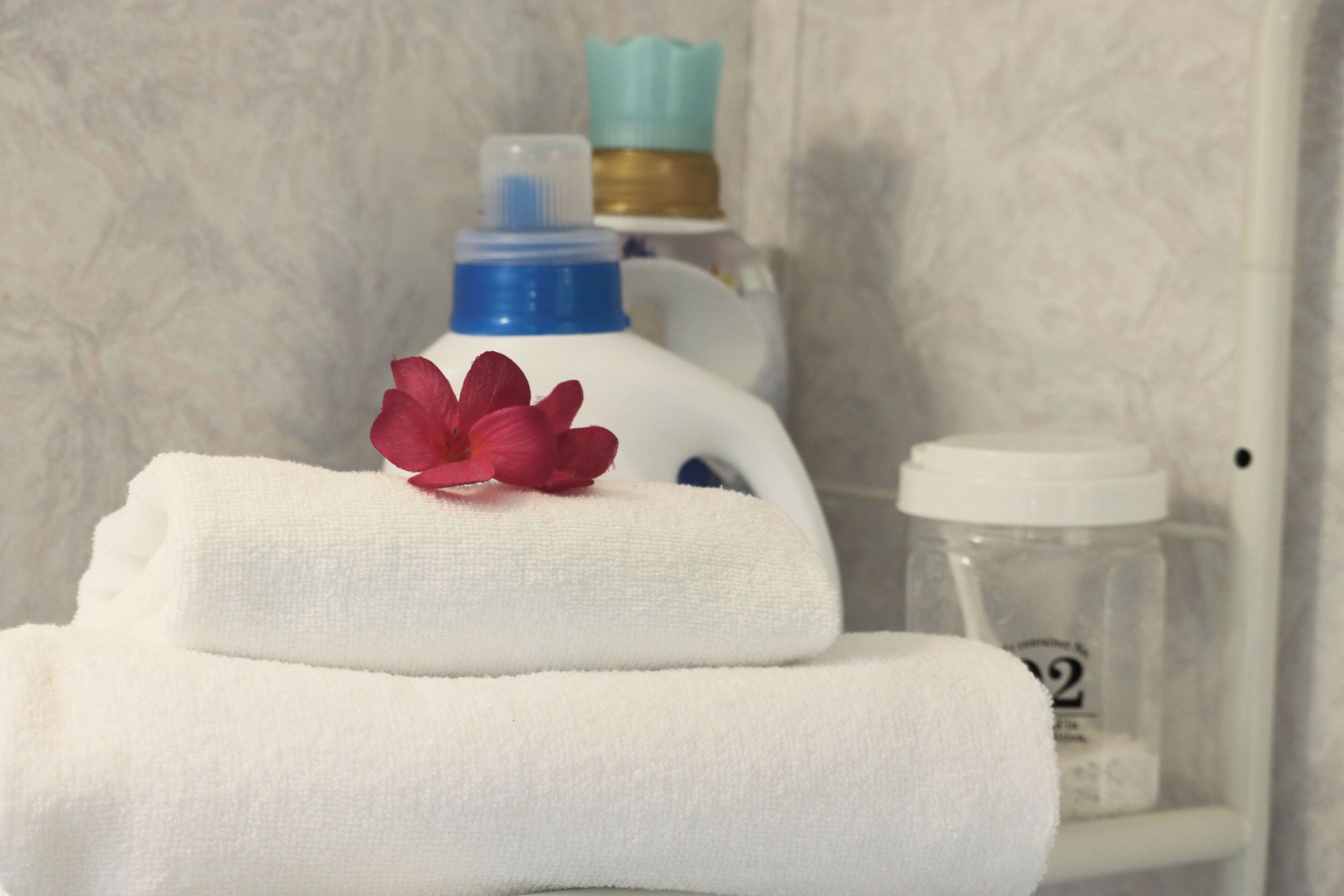 Une pile de serviettes blanches surmontée d’une fleur rose, une bouteille bleue et blanche, et un bocal transparent avec un couvercle blanc en arrière-plan.
