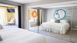 Et hotelværelse med et stort spejl, der viser en seng, en stol og et solmaleri på væggen.