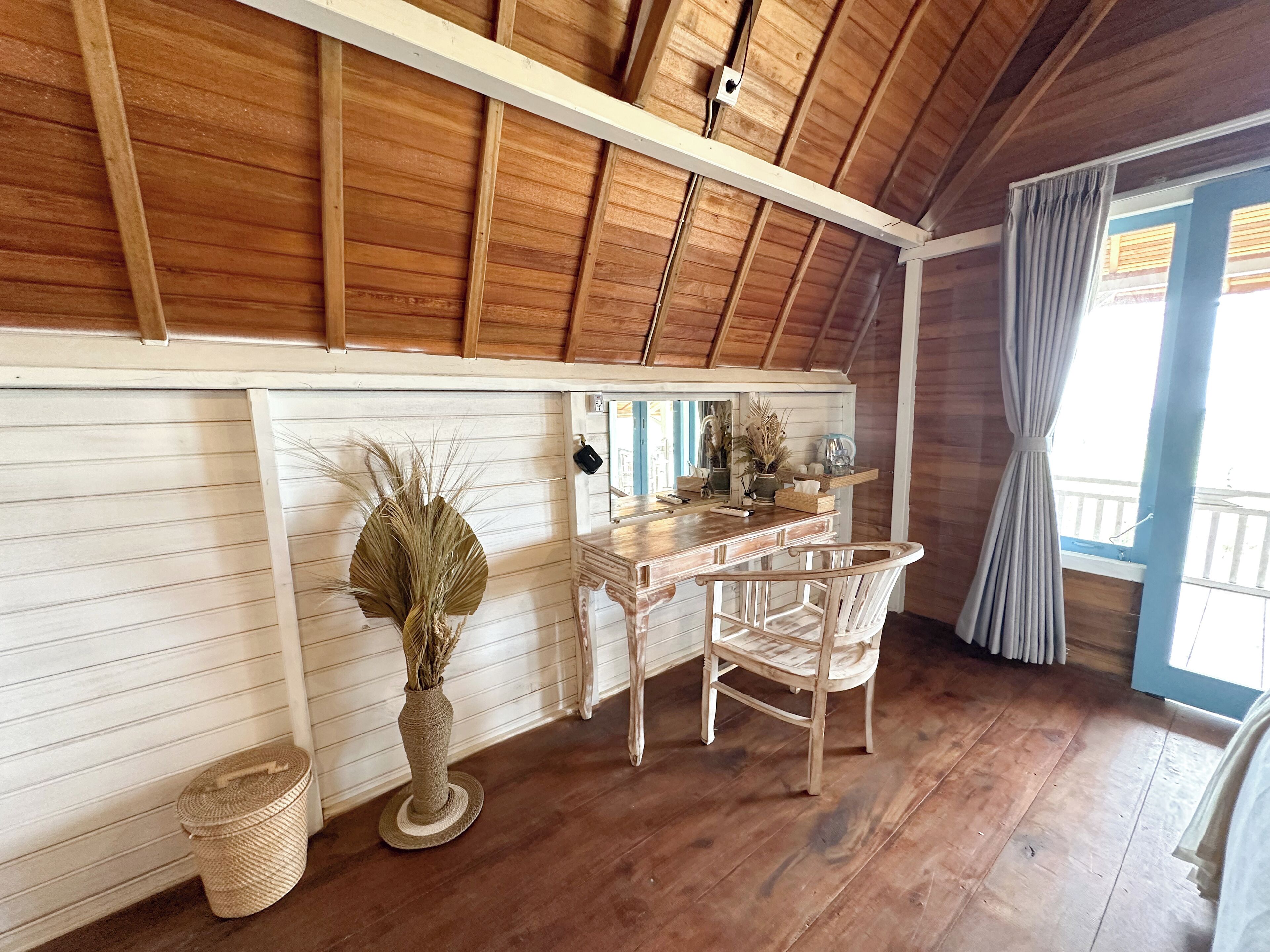 A rustic kitchen with wooden beams, a wooden counter, a chair, a vase with dried plants, and a window with curtains.
