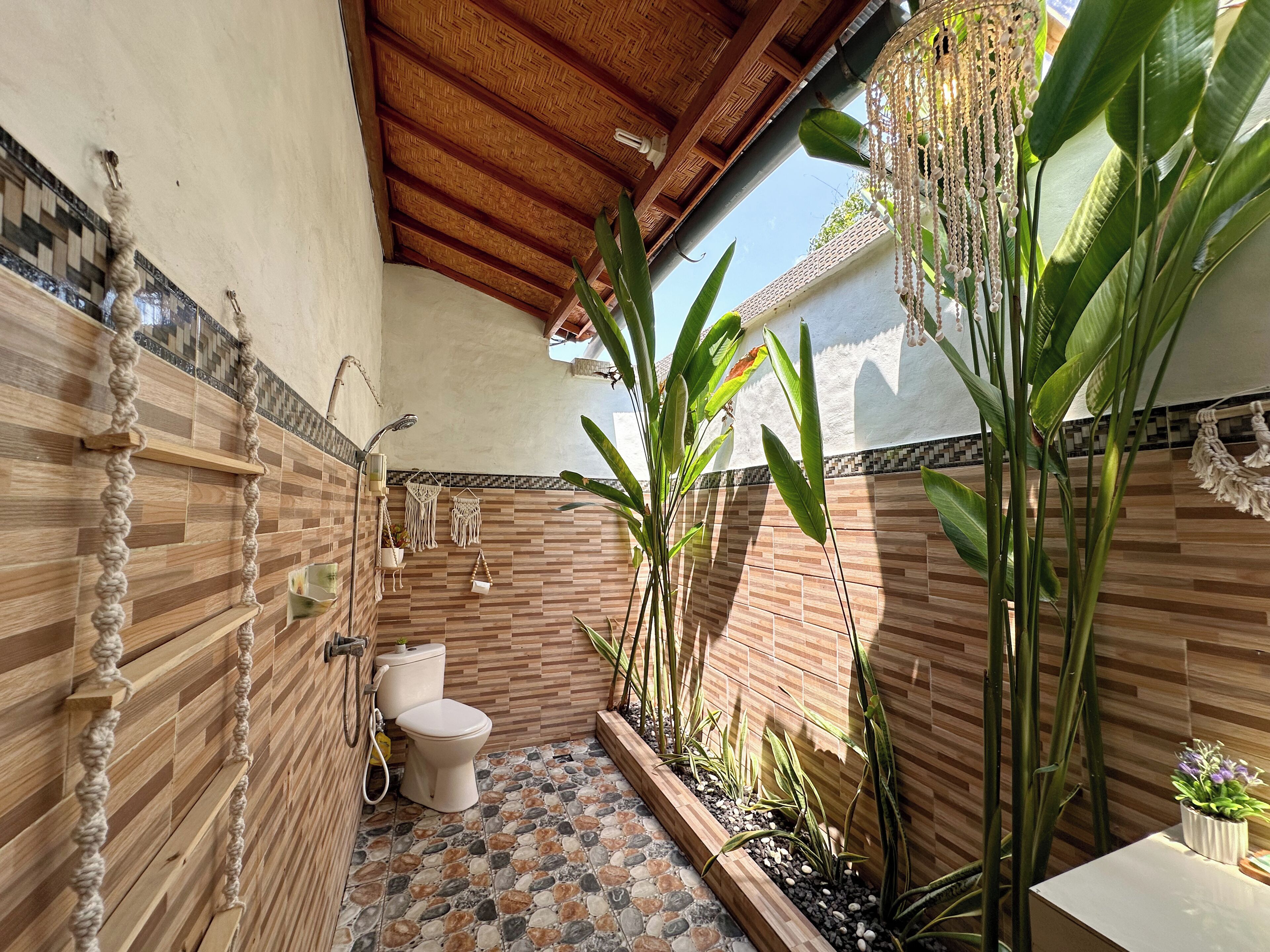 A bathroom with a toilet, a shower, and a sink. It has a tiled floor and walls with a wooden ceiling. There are plants and a hanging macrame decoration.