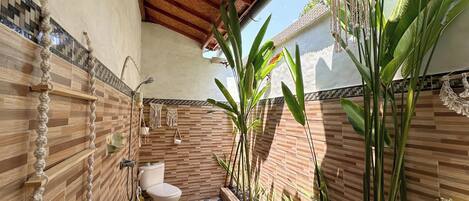 A bathroom with a toilet, a shower, and a sink. It has a tiled floor and walls with a wooden ceiling. There are plants and a hanging macrame decoration.