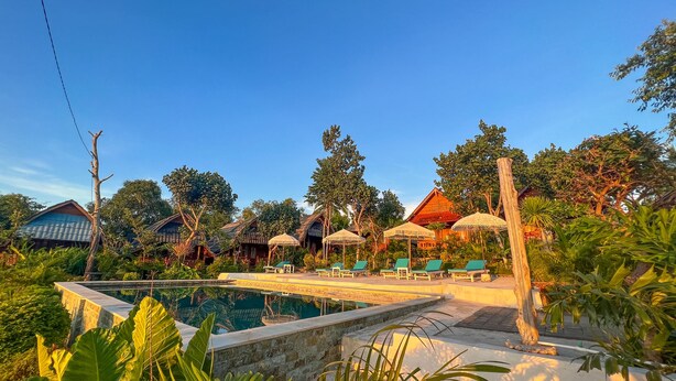 A pool area with lounge chairs and umbrellas, surrounded by lush greenery and traditional-style buildings. - Sunset Hill Penida Resort (Penida Island)