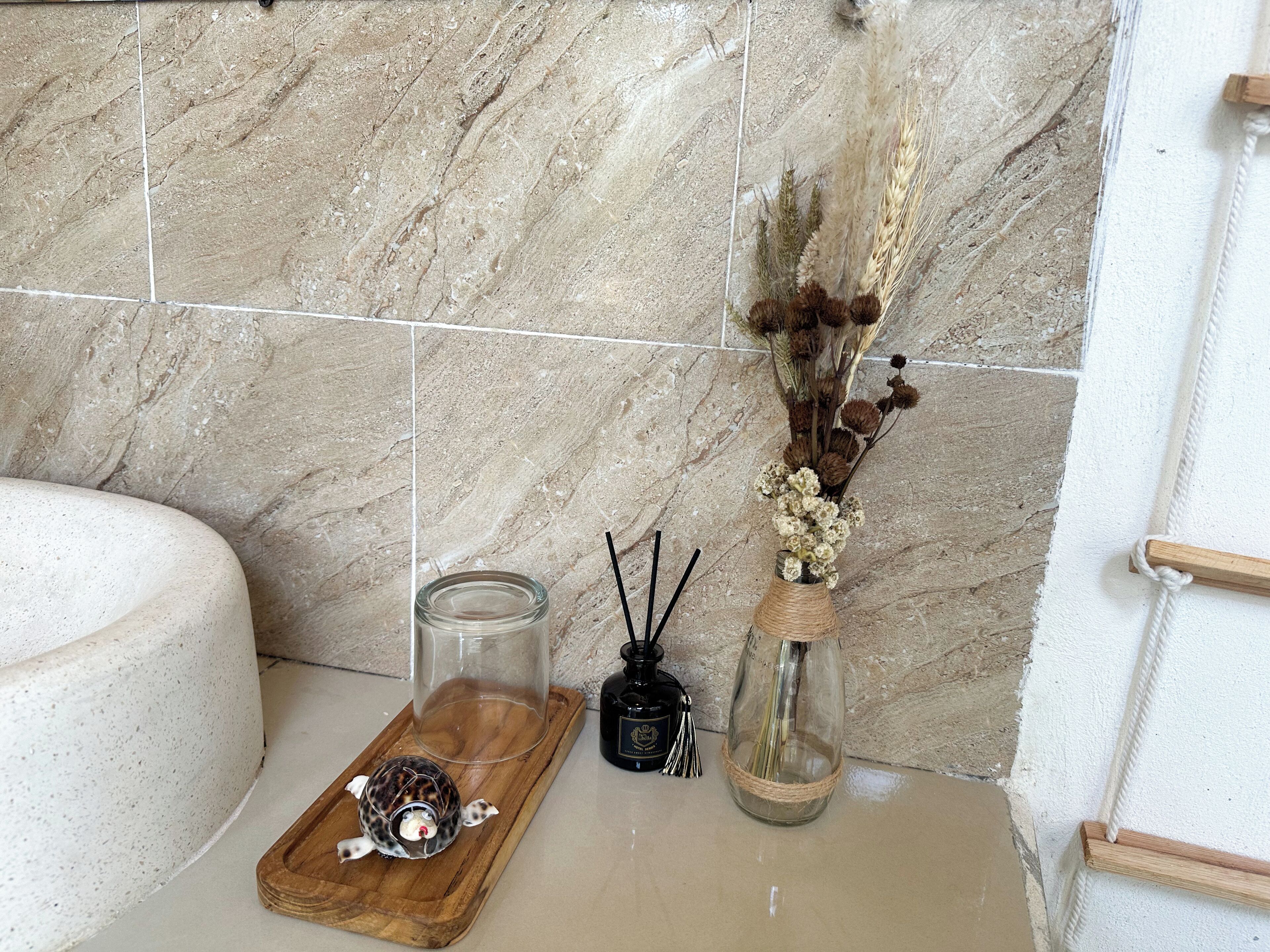 A bathroom with a marble wall, a wooden tray with a decorative item, a glass jar, and a vase with dried flowers.