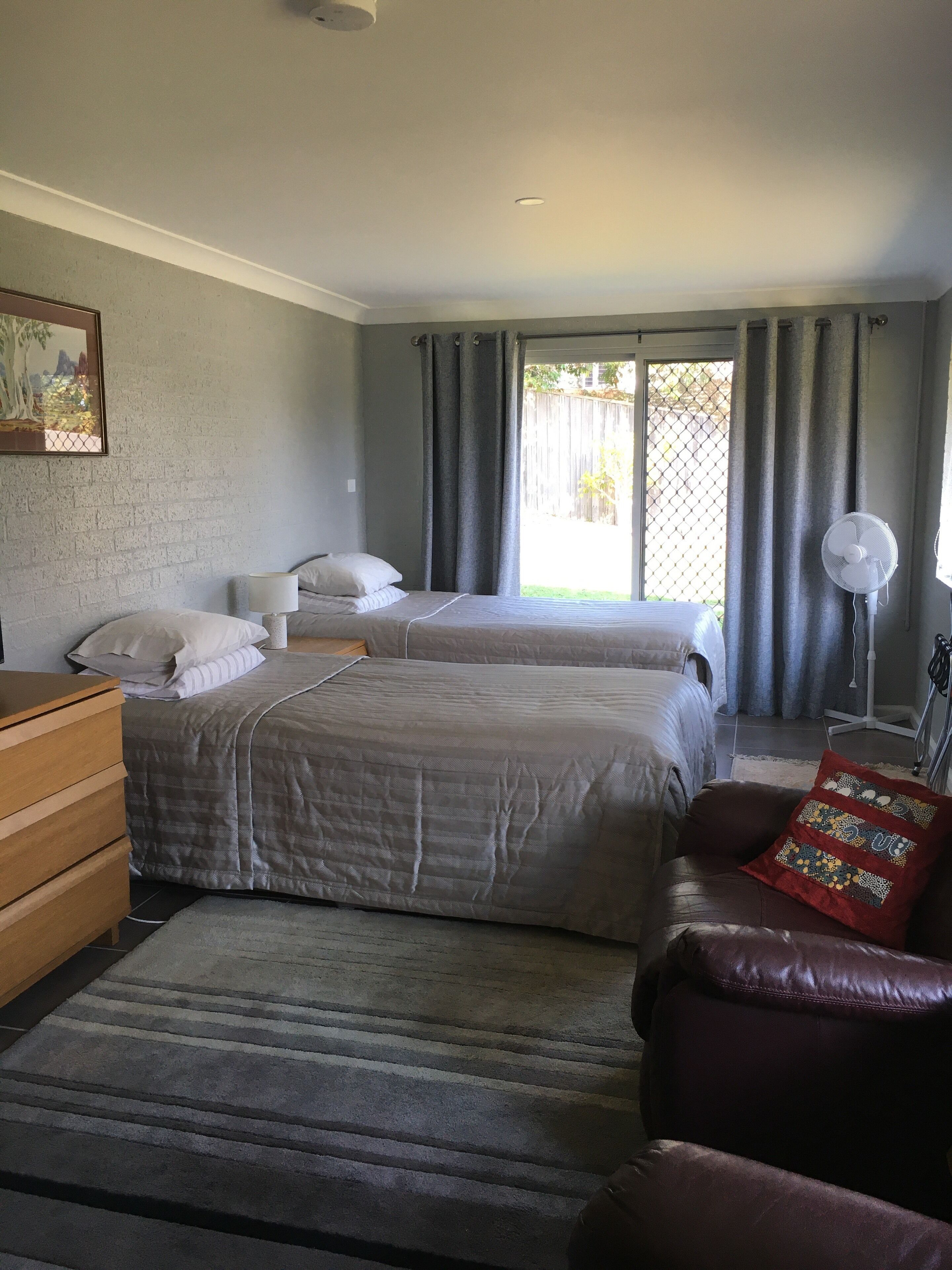A bedroom with two beds, a wooden dresser, a red cushioned chair, a standing fan, and a window with curtains.