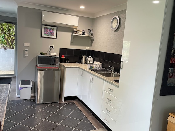 A modern kitchen with white cabinets, a stainless steel refrigerator, a microwave, and a kettle. There is a black and white backsplash, a clock on the wall, and a window with a view of greenery outside.