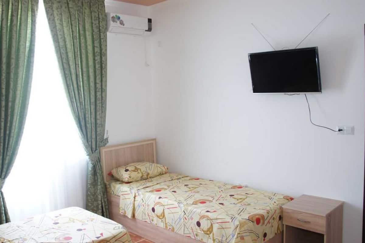 A small, single-room hotel room with a bed, a bedside table, a TV mounted on the wall, and a window with curtains.