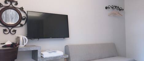 A modern hotel room with a flat-screen TV, a sofa, a small table with a kettle, and a trash bin.