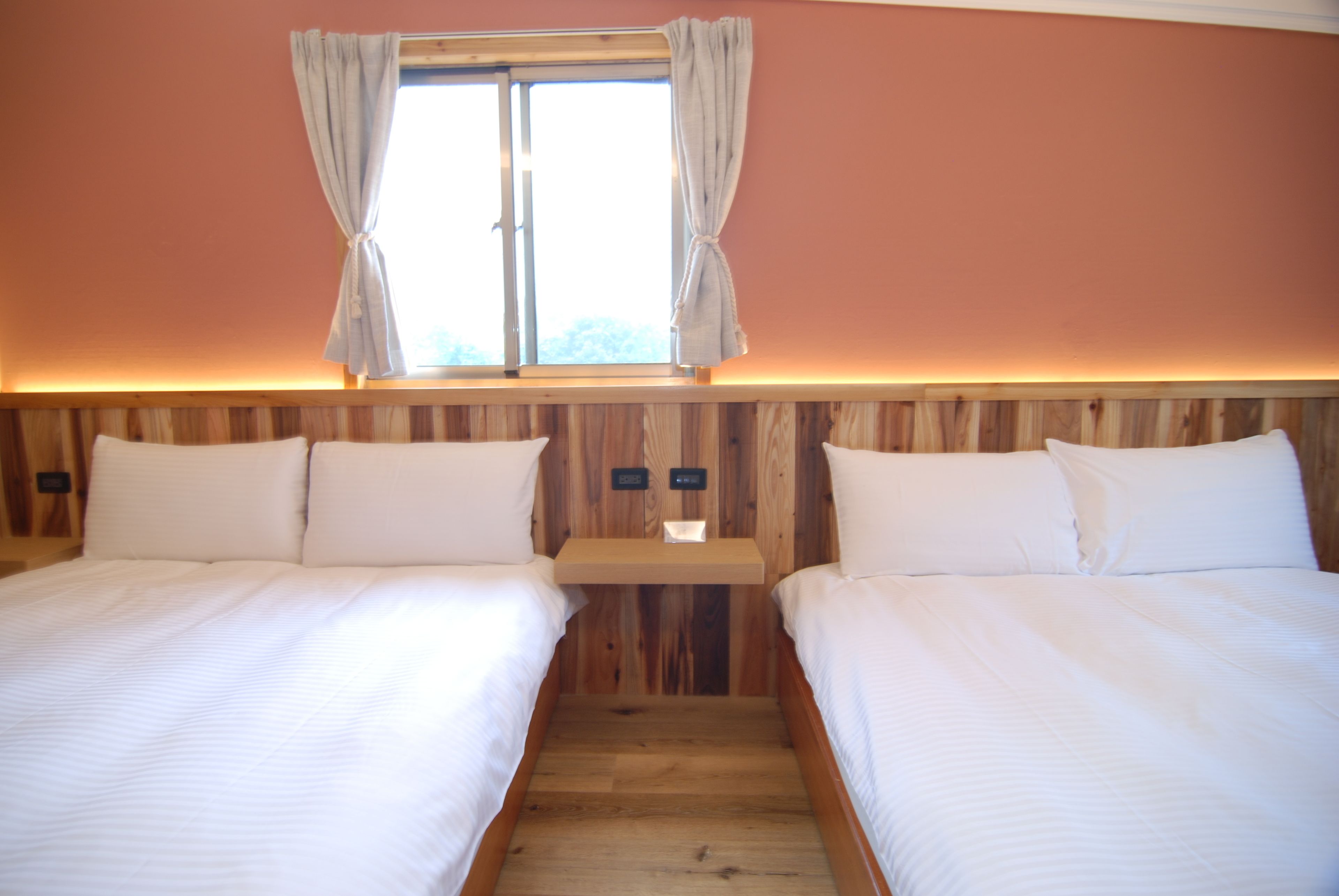 Une chambre avec deux lits, une tête de lit en bois, une table de chevet avec une lampe et une fenêtre avec des rideaux.