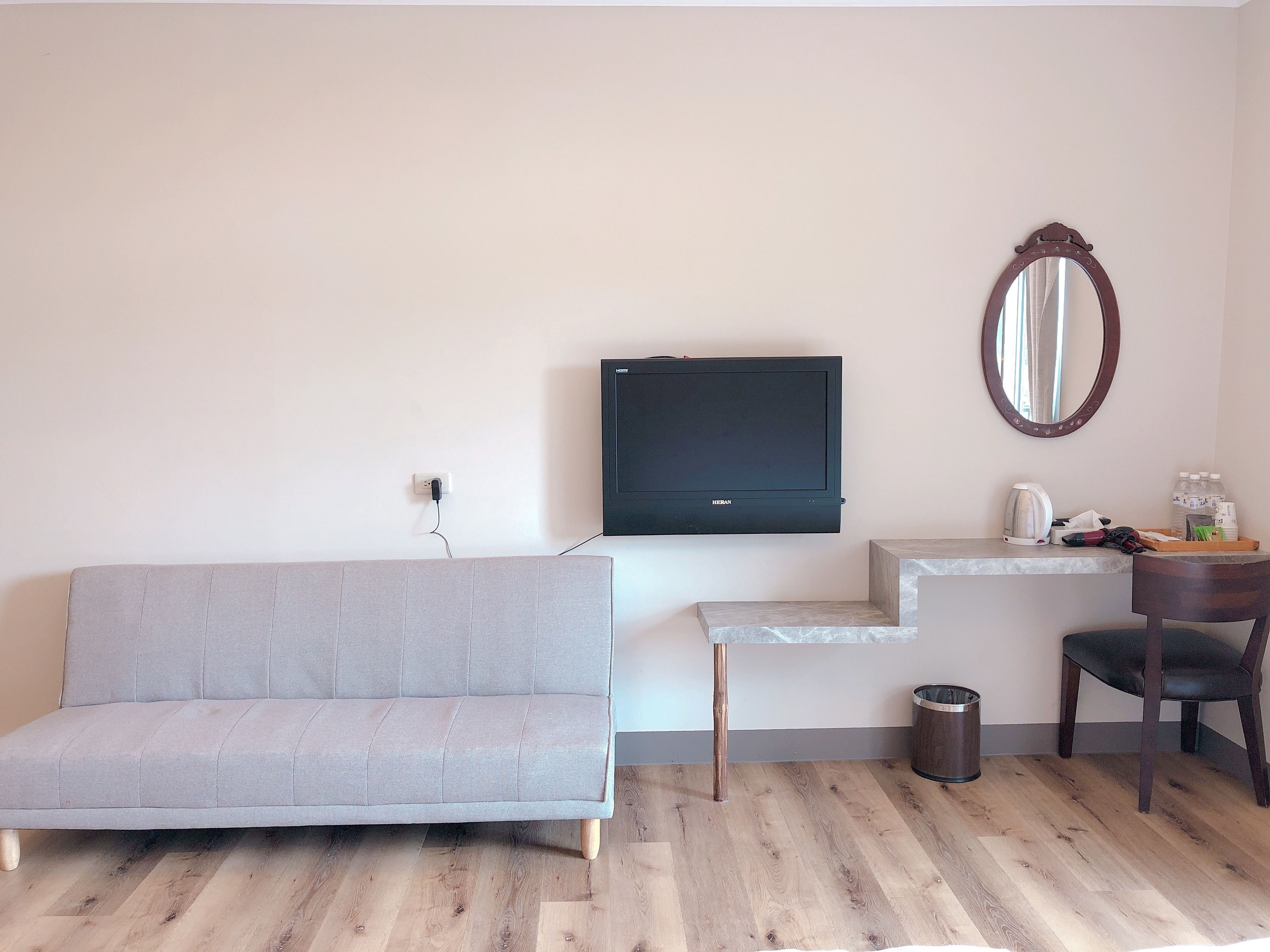 A modern living room with a grey sofa, a mounted flat-screen TV, a wooden coffee table, a small trash bin, and a wooden chair.