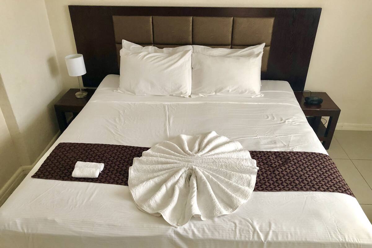 Un lit bien fait, avec du linge de lit blanc, une tête de lit marron et un linge décoratif disposé au centre.