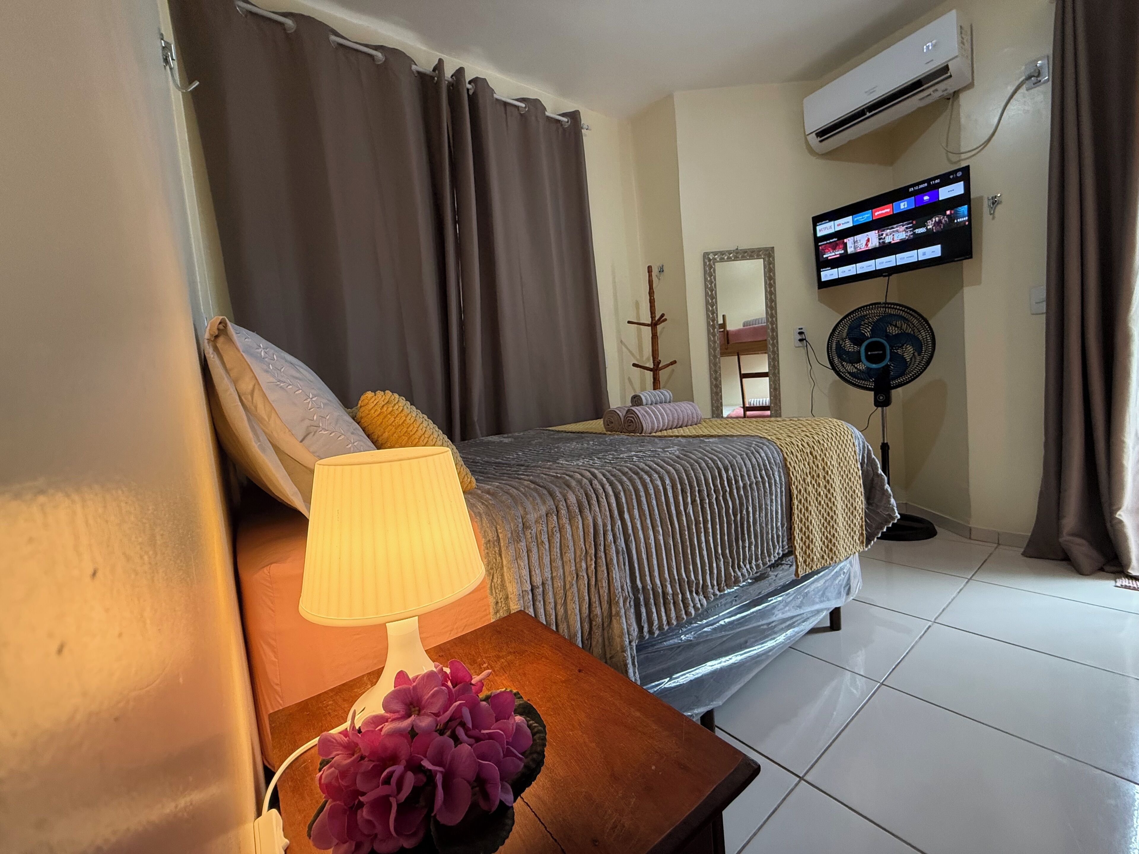 A hotel room with a bed, a bedside table with a lamp and flowers, a TV on the wall, and an air conditioner.