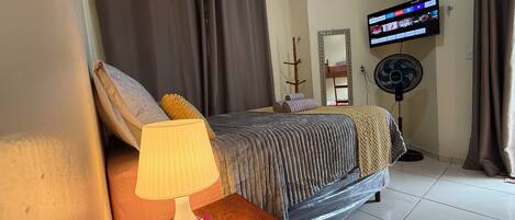 A hotel room with a bed, a bedside table with a lamp and flowers, a TV on the wall, and an air conditioner.